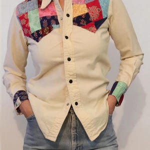 May include: A cream colored long-sleeved button-up shirt with a patchwork design on the yoke. The patchwork is made of various colors and patterns, including red, blue, green, and yellow.