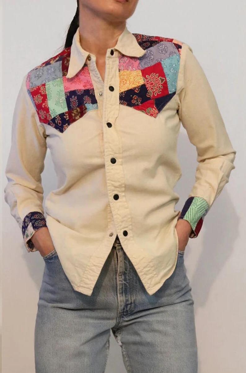 May include: A cream colored long-sleeved button-up shirt with a patchwork design on the yoke. The patchwork is made of various colors and patterns, including red, blue, green, and yellow.