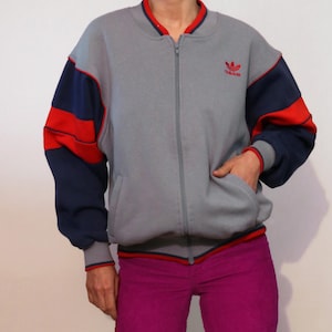 Op de afbeelding: Een grijs jack met rits met rood-blauwe kleurblokarmen. Het jack heeft een rood-blauwe bies rond de kraag en de manchetten. Het Adidas-logo is in rood geborduurd op de linkerborst.