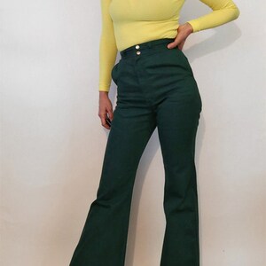 Puede incluir: Una mujer lleva un jersey de cuello alto amarillo brillante y pantalones anchos de color verde oscuro con cintura alta y cierre de bot&oacute;n.