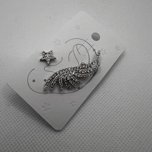 May include: A pair of silver earrings with a star and a feather design. Both pieces are encrusted with clear rhinestones.