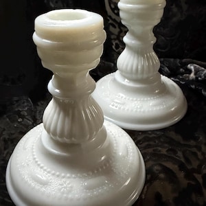 May include: Two white milk glass candle holders. The candle holders have a round base with a detailed pattern, a fluted stem, and a circular top. The candle holders are made of opaque white glass and have a vintage aesthetic.