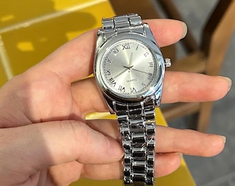 May include: Silver wristwatch with a round face and Roman numeral hour markers. The watch has a silver band and a silver dial. The watch is held against a yellow tiled surface.