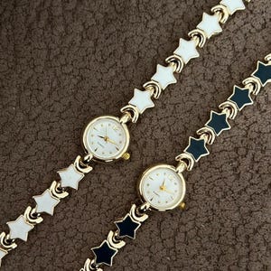 May include: Two gold-toned wristwatches with star-shaped links. One watch has white links, the other has black links. Both watches have a round, gold-colored watch face with gold hands and markers. The watches are on a brown surface.