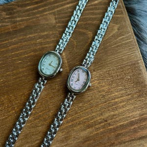 May include: Two silver oval-shaped wristwatches with metal bands. One watch has a white face with black Roman numeral markings and the other has a pink face with black Roman numeral markings. Both watches say "QUARTZ" on the face.