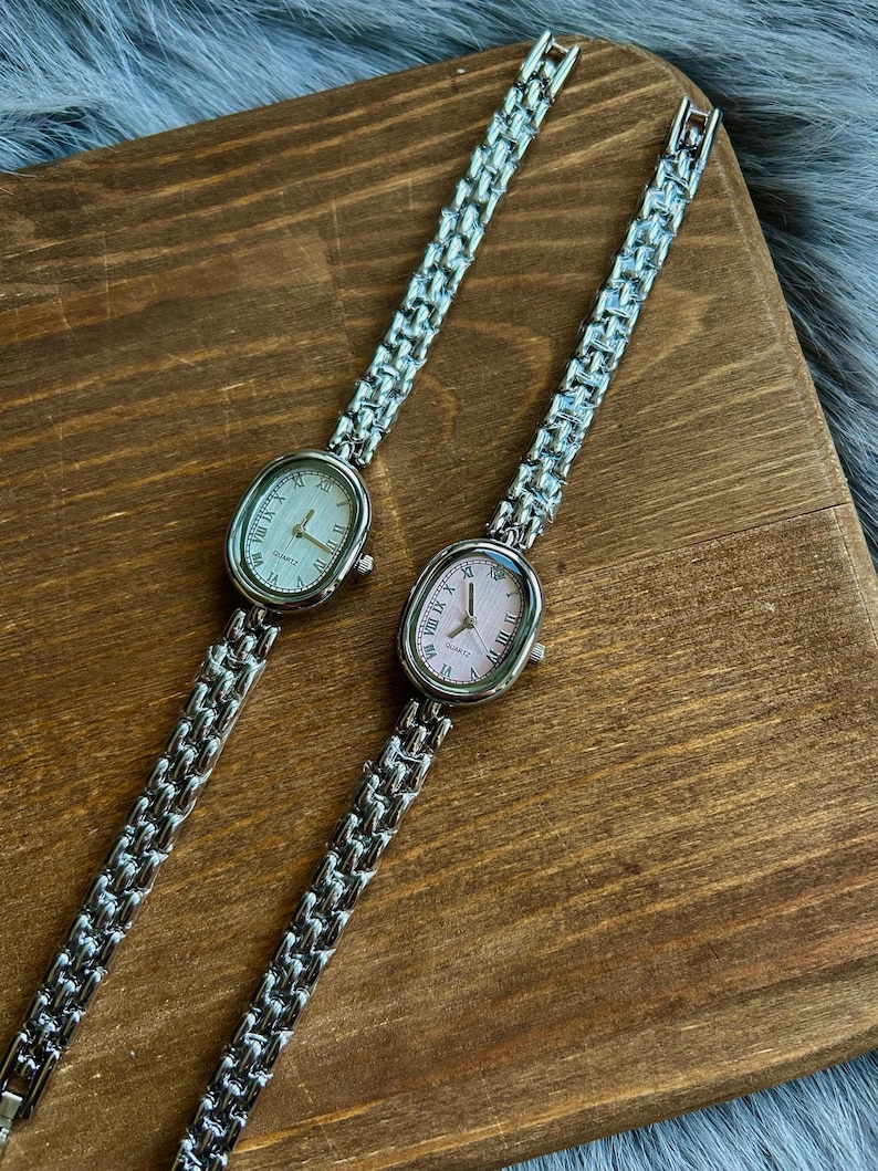 May include: Two silver oval-shaped wristwatches with metal bands. One watch has a white face with black Roman numeral markings and the other has a pink face with black Roman numeral markings. Both watches say "QUARTZ" on the face.