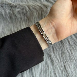 May include: A silver chain bracelet with a clasp closure. The bracelet is on a person's wrist.