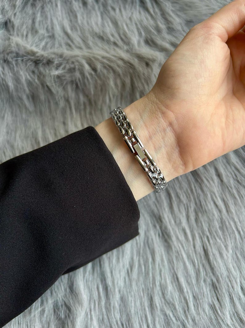May include: A silver chain bracelet with a clasp closure. The bracelet is on a person's wrist.