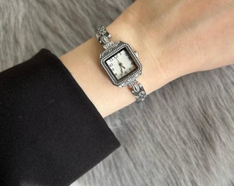 May include: Silver-toned wristwatch with a square face and a white dial. The watch has a decorative band with floral accents. The watch is worn on a wrist, with a black sleeve visible in the background.