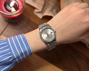 May include: Silver wristwatch with a round, light-colored face and a silver band. The watch has a bezel with small, clear stones. The watch is on a wrist with a blue and white striped sleeve.