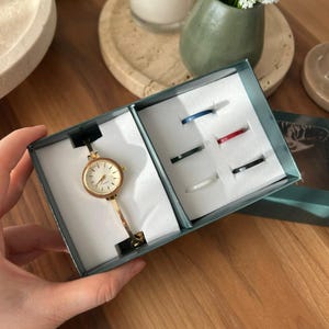 Op de afbeelding: Een goudkleurig polshorloge met een ronde wijzerplaat en bijpassende armband, gepresenteerd in een turquoise geschenkdoos. De doos bevat ook een set van zes kleurrijke ringen. De ringen zijn in verschillende kleuren, waaronder blauw, rood en zwart.