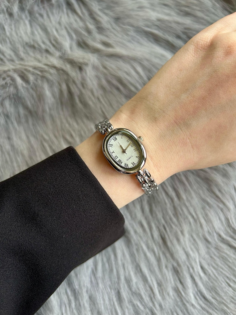 May include: A silver oval-shaped watch with Roman numeral markings on the face. The watch has a silver metal band.