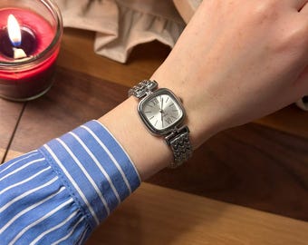 May include: A silver-toned wristwatch with a square face and Roman numeral hour markers. The watch has a linked band and is worn on a wrist with a blue and white striped shirt sleeve.