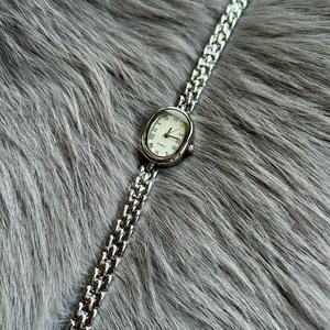 May include: A silver watch with a white face and Roman numeral hour markers. The watch has a silver metal band.