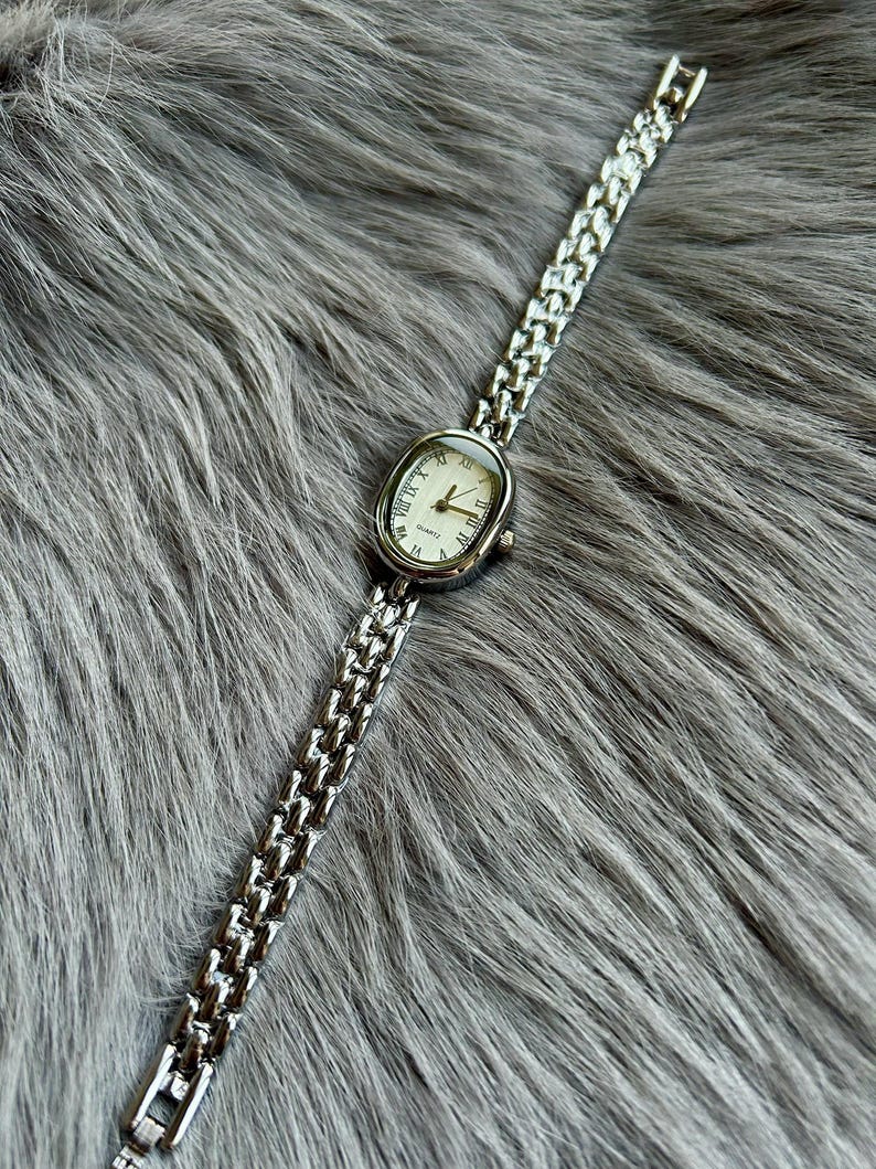 May include: A silver watch with a white face and Roman numeral hour markers. The watch has a silver metal band.