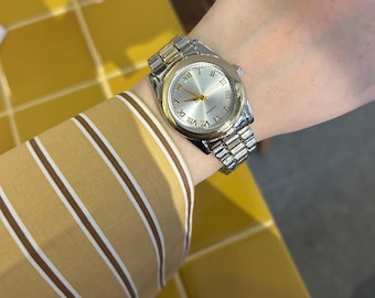 May include: A silver and gold-toned quartz watch with a silver face and gold hands and Roman numeral hour markers. The watch is on a silver and gold-toned metal band. The watch is being worn on a wrist.