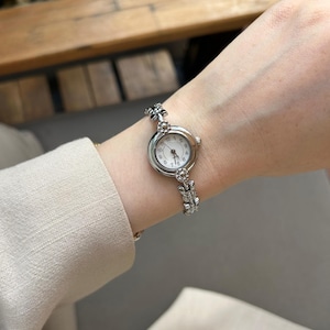 Puede incluir: Reloj de pulsera plateado con esfera blanca y números romanos. La pulsera decorativa es un elemento clave. El reloj se lleva en la muñeca, con una manga de tono neutro en el fondo.