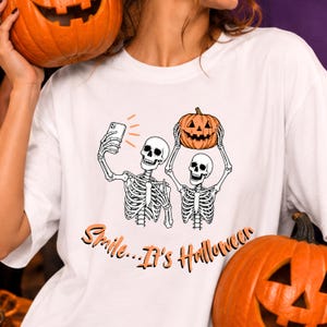 Puede incluir: Una camiseta blanca con un gráfico de dos esqueletos tomándose un selfie con un smartphone mientras uno sostiene una calabaza naranja tallada sobre su cabeza. Debajo de los esqueletos, el texto "Smile...It's Halloween" aparece en letras naranjas.