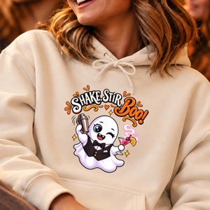 Puede incluir: Una sudadera con capucha de color crema que presenta un gráfico de un fantasma blanco con chaleco negro y pajarita. El fantasma sostiene una coctelera y una bebida rosa con una rodaja de naranja. El texto superior dice SHAKE, STIR, BOO!.