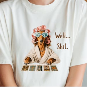 Puede incluir: Una camiseta blanca con el gráfico de un perro marrón que lleva rulos rosas, gafas de sol en forma de corazón y un albornoz beige. El perro tiene un cigarrillo en la boca y apoya las patas sobre cinco cartas del tarot. El texto dice Well... Shit.