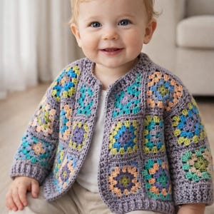 May include: A baby wearing a handmade, crocheted cardigan. The cardigan is a patchwork of colorful squares in shades of blue, green, purple, and peach, set against a gray background. The baby is also wearing a white shirt and khaki pants.