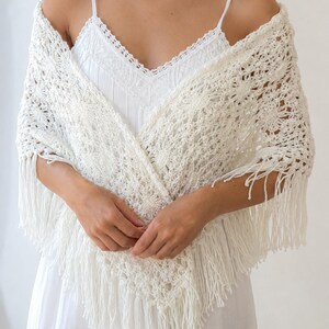 May include: A white triangular crochet shawl featuring an intricate openwork pattern and long fringe along the edges. The shawl is draped over a white camisole with thin straps and a lace-trimmed V-neckline, creating a layered, textured appearance.