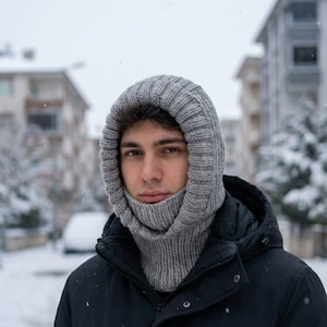 Può includere: Un passamontagna grigio lavorato a maglia, che copre la testa e il collo, è indossato sopra una giacca nera. Il passamontagna ha una trama a coste e una vestibilità aderente. Lo sfondo mostra una scena esterna innevata con edifici e alberi.