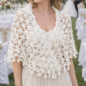 May include: A white crochet shawl featuring an intricate openwork star pattern with a V-neckline and a scalloped hem finished with small decorative tassels. The garment is crafted from a soft yarn and drapes over the shoulders and upper torso.