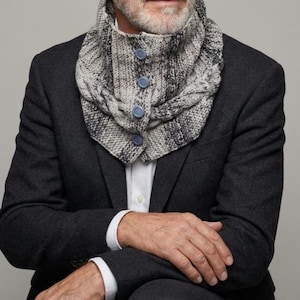 May include: A grey and white marled knit cowl scarf featuring a thick cable knit texture and a vertical row of four round blue buttons down the center front. The scarf is worn over a white collared shirt and a dark charcoal grey blazer jacket.
