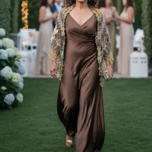 May include: A floor-length brown satin dress with a V-neckline and a sleeveless bodice, paired with a multi-colored crochet shawl featuring intricate floral patterns and fringe edges. A small metallic clutch is held in one hand.