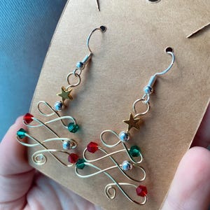 May include: A pair of handmade Christmas tree earrings. The earrings feature a gold-colored wire design with red and green beads, silver beads, and a gold star at the top. The earrings are displayed on a brown cardboard backing.