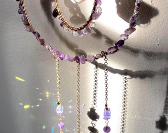 May include: A hanging ornament featuring two concentric gold wire hoops wrapped in purple amethyst chips. A raw amethyst crystal hangs in the center. Four gold chains dangle from the bottom, adorned with small amethyst beads and a clear glass prism.
