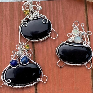 May include: Three wire-wrapped pendants featuring large, smooth, oval black stones. Each pendant is encased in intricate silver-toned wire coils. One pendant includes two small blue stones, another features two small iridescent stones, and all have tiny gold accents.