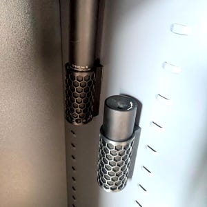 May include: Two black metal suppressor mounts with a honeycomb pattern. The mounts are attached to a metal wall.