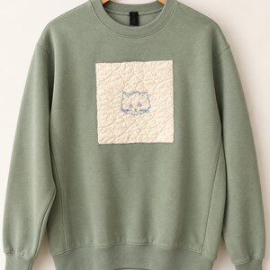 Könnte beinhalten: Ein salbeigrünes Sweatshirt mit Rundhalsausschnitt und einem quadratischen, cremefarbenen Steppaufnäher in der Mitte der Brust. Der Aufnäher zeigt eine einfache, dunkelblau gestickte Linienzeichnung eines Katzengesichts.