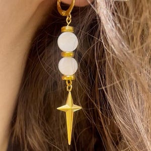 Könnte beinhalten: Goldfarbene Creolen mit einem hängenden Design. Jeder Ohrring hat zwei weiße, runde Perlen, die durch goldfarbene Akzente getrennt sind, und einen goldenen Sternanhänger am unteren Ende. Die Ohrringe werden vor einem Hintergrund aus braunem Haar gezeigt.