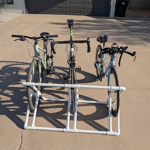 May include: Three bicycles of different styles are displayed on a white PVC pipe bike rack. The bikes are a light green mountain bike, a black road bike, and a blue and black road bike. The bikes are parked on a concrete driveway.