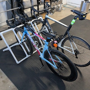 May include: Two road bikes are displayed on a white PVC pipe stand. One bike is light blue with red and purple accents, the other is black and white. The bikes are on a black rubber mat in a garage setting.