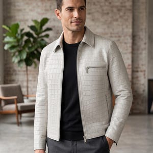 Puede incluir: Una chaqueta gris claro con un patrón texturizado de relieve de cocodrilo, cuello en punta y cremallera frontal completa. La chaqueta cuenta con un bolsillo horizontal con cremallera en el pecho y dos bolsillos laterales, sobre una camisa negra y pantalones gris oscuro.