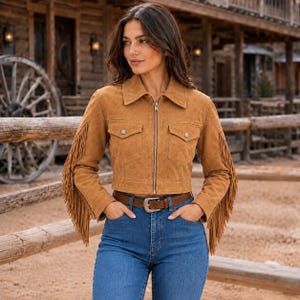 Puede incluir: Una chaqueta de ante color tostado con cremallera frontal, cuello en punta y dos bolsillos en el pecho con solapas abotonadas. Las mangas están adornadas con largos flecos colgantes. La chaqueta se lleva con vaqueros azules y un cinturón marrón.