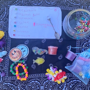 Puede incluir: Una colección de accesorios y materiales de manualidades coloridos, que incluyen coleteros, pegatinas, pulseras y slime. Una lista escrita a mano detalla artículos como llaveros y gemas de cristal. Un tarro contiene cuentas.
