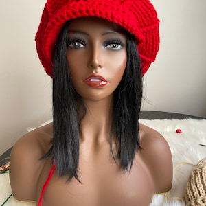 May include: A bright red crocheted newsboy cap featuring a thick, textured yarn construction with a prominent brim. The hat has a rounded, structured crown with visible vertical stitching patterns throughout the fabric, creating a distinct ribbed appearance.
