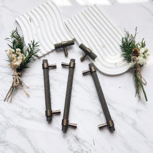 May include: A collection of bronze-toned cabinet pulls and knobs. The pulls have a textured, cylindrical design with a T-shaped handle. The knobs are shorter with a similar textured finish. The hardware is arranged on a white surface with decorative elements.