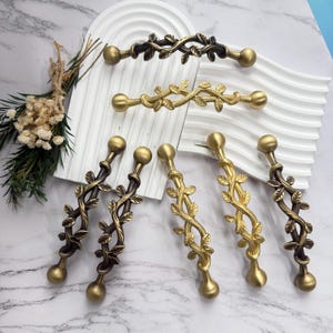 May include: A collection of decorative cabinet pulls and handles in bronze and gold tones. The hardware features a leaf and vine design, with a ball-shaped end on each piece. The pulls are arranged on a white surface.