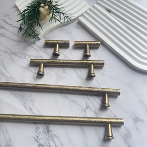 May include: A collection of antique brass cabinet hardware, including handles and knobs. The hardware features a hammered texture and a warm, golden hue. The knobs are T-shaped, and the handles are bar-shaped, varying in length.