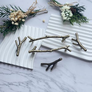 May include: A collection of bronze-colored branch-shaped cabinet pulls and knobs. The hardware is arranged on white, textured trays, with dried floral arrangements in the background. The pulls and knobs have a rustic, natural aesthetic.