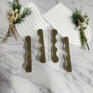 May include: Four antique brass cabinet pulls with a wavy design are arranged on a marble surface. The pulls are a warm, metallic color. Decorative white, wave-shaped trays and dried floral arrangements are in the background.