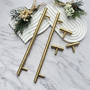 May include: A set of gold-colored cabinet hardware, including two long bar pulls and four T-shaped knobs. The hardware has a hammered texture and is displayed on a white surface with decorative floral accents.
