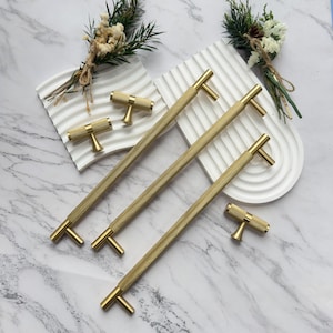 May include: Gold-toned cabinet hardware set. The set includes long bar pulls and smaller T-shaped knobs. The hardware has a textured, ridged design on the bar and knob handles. The hardware is displayed on a white surface with decorative greenery.