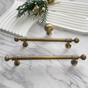 May include: Antique brass cabinet hardware set. Includes two long pull handles and a round knob. The handles have a ribbed ball design on each end. The hardware is displayed on a white tray and marble surface.